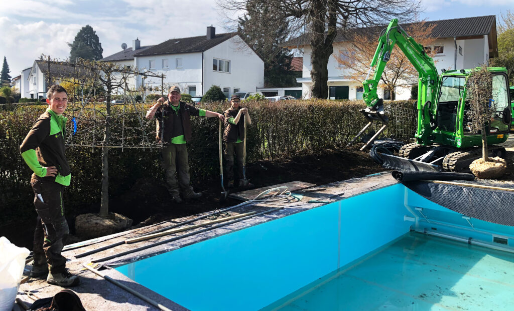 Zeigt Mitarbeiter vor Pool im Fertigstellung. Im Hintergrund ein Bagger mit Palettenschaufel zum Baumtransport