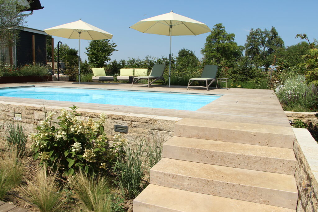 Das Bild zeigt einen Pool umgeben mit feinem Terrassensteinbelag, Bepflanzung, Sitzen und Sonnenschirmen.